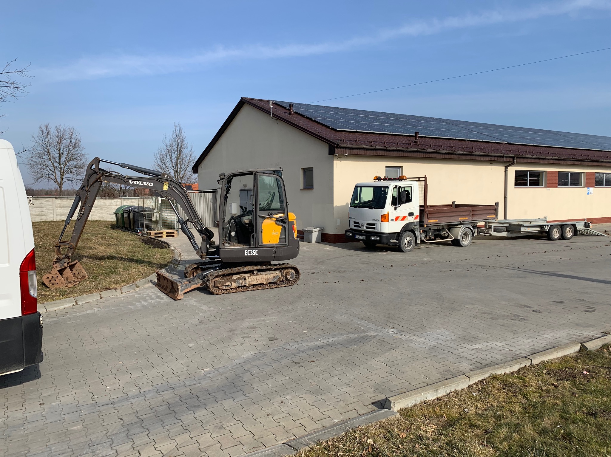 Mini koparka Volvo EC35C na kostce brukowej, obok ciężarówka z przyczepą, budynek z panelami słonecznymi w tle. Dzień.