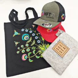 Czarny bawełniany worek z kolorowym folkowym wzorem kwiatowym, dwie czapki z daszkiem (zielona i czerwona) z logo HFT71 oraz szara filcowa kosmetyczka z napisem SEE SEA, leżące na białym tle.