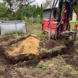Czerwona wiertnica na gąsienicach stoi obok wykopu z pryzmą piasku, w tle zbiornik na wodę i drzewa.