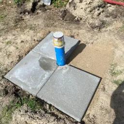 Nisko osadzona studnia z niebieską rurą wystającą ponad powierzchnię, otoczona czterema betonowymi płytami chodnikowymi, z widoczną łopatą i poziomicą w tle.