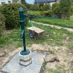 Studnie Głębinowe Paweł Moryto - Zielona pompa ręczna zamontowana na betonowej podstawie, obok taczka z piaskiem i narzędzia, na tle domów jednorodzinnych i zieleni.