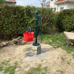 Zainstalowana zielona pompa ręczna z czerwonym wiadrem na betonowej podstawie, otoczona piaskiem i trawą, z żywopłotem w tle.