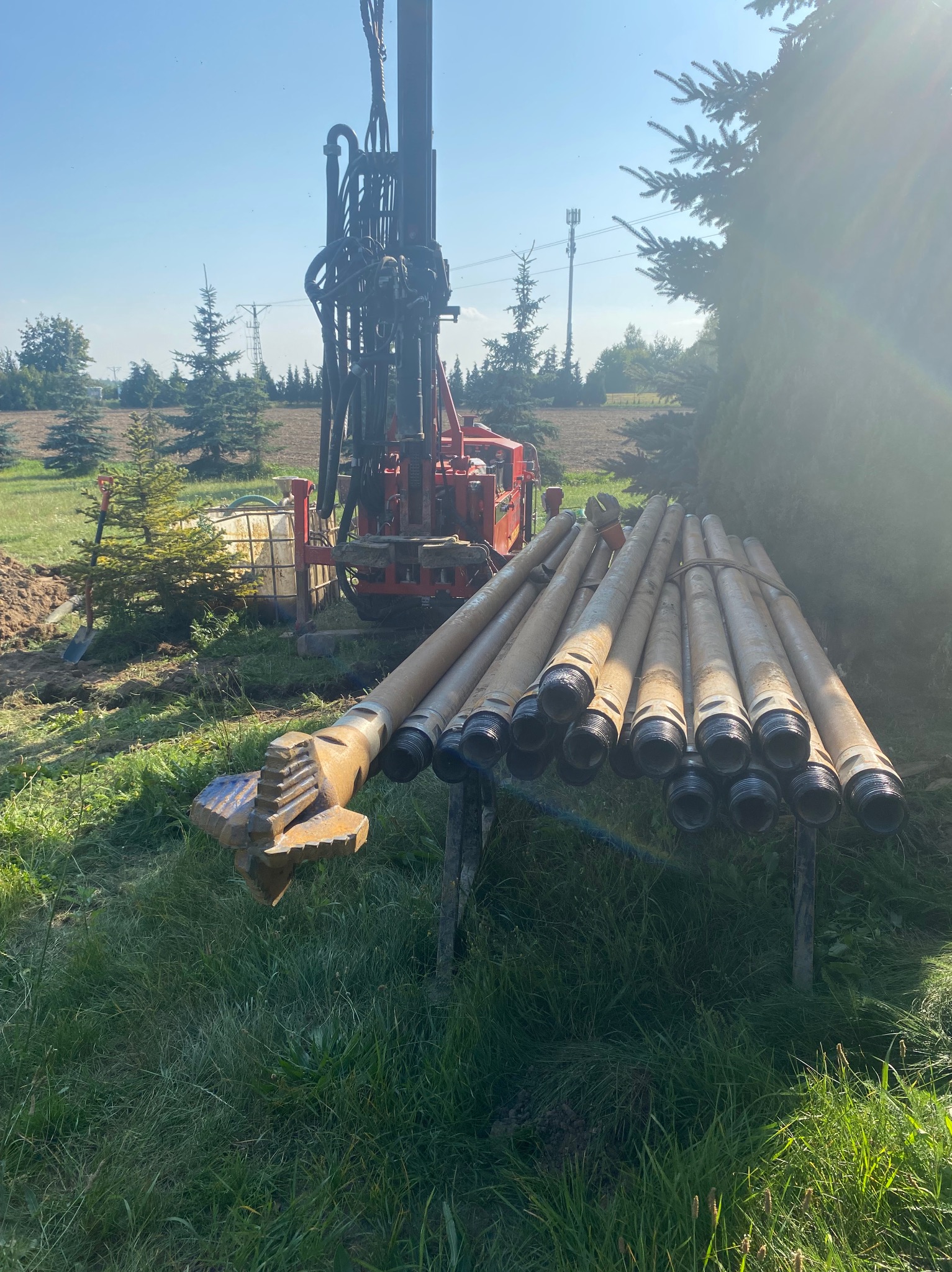 Wiertnica do studni głębinowych na tle zieleni, z rurami wiertniczymi ułożonymi na stojaku i charakterystycznym wiertłem z przodu.