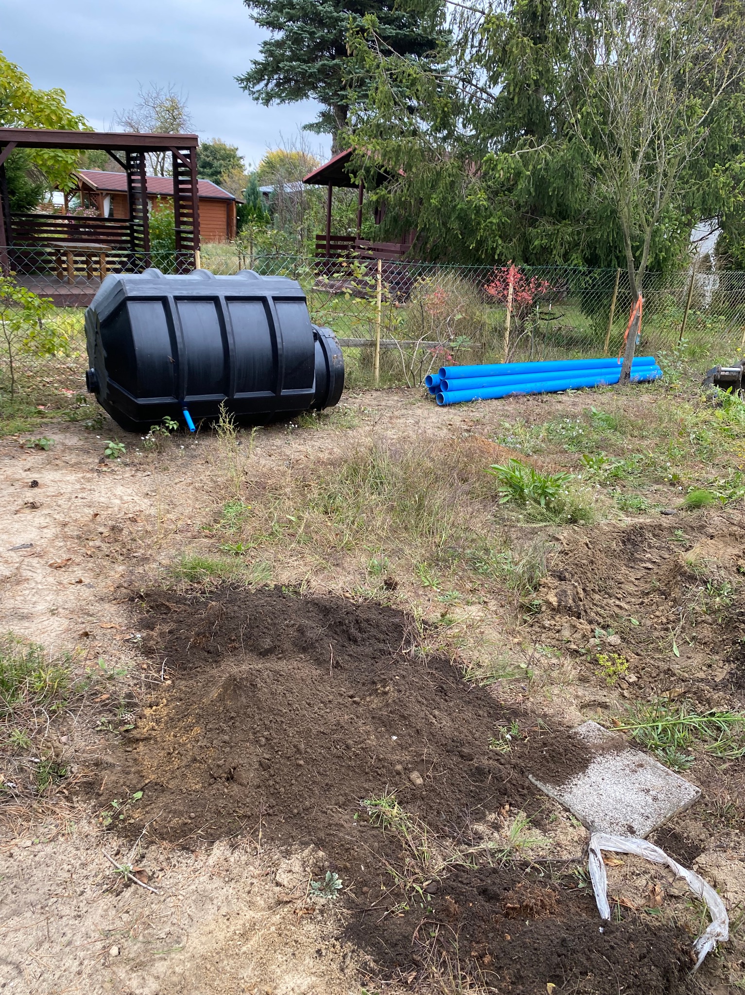 Czarny zbiornik retencyjny na wodę deszczową leżący obok wykopu w ziemi, z rurami PCV w tle i drewnianą altaną w oddali.
