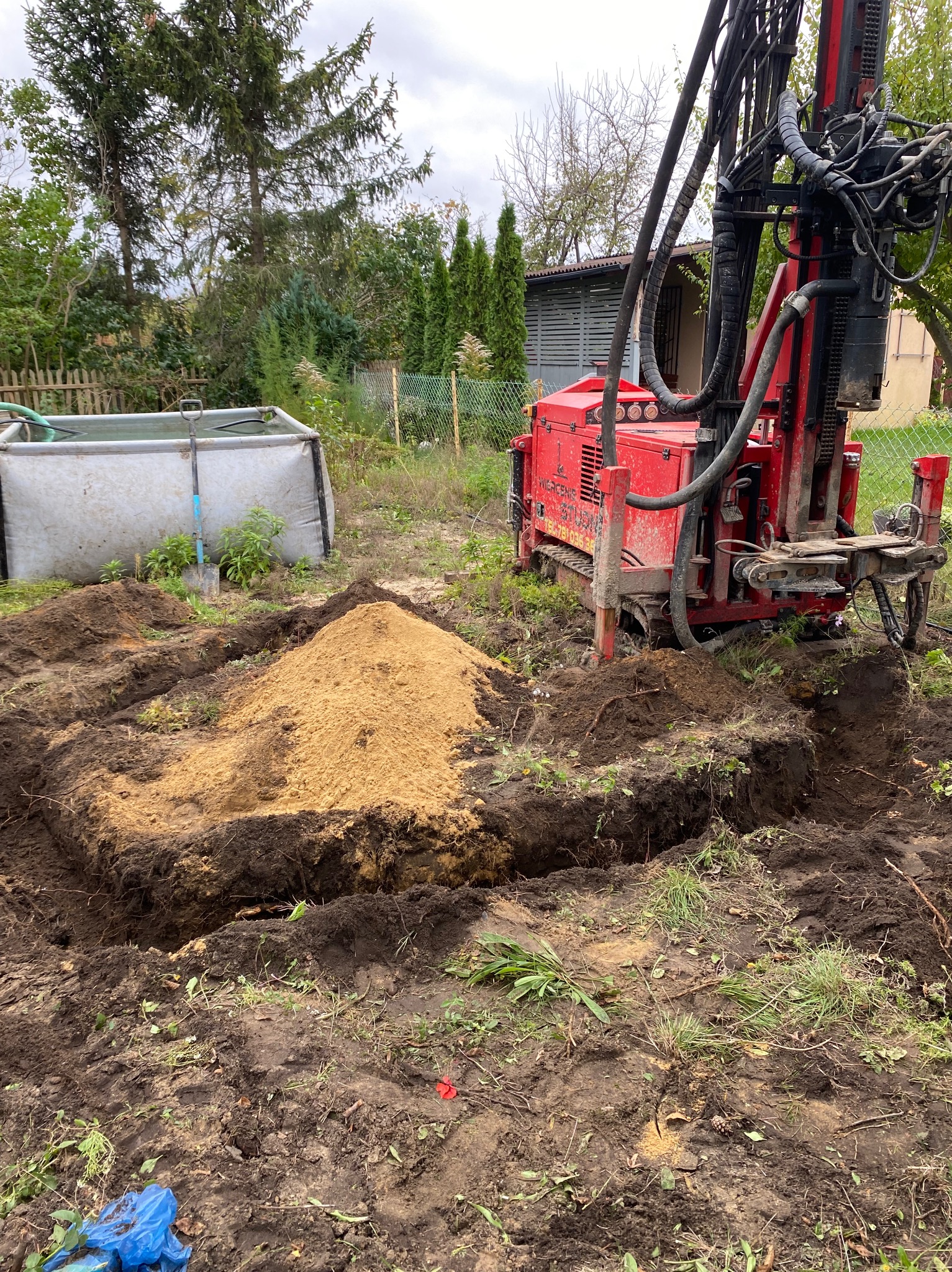 Czerwona wiertnica na gąsienicach stoi obok wykopu z pryzmą piasku, w tle zbiornik na wodę i drzewa.