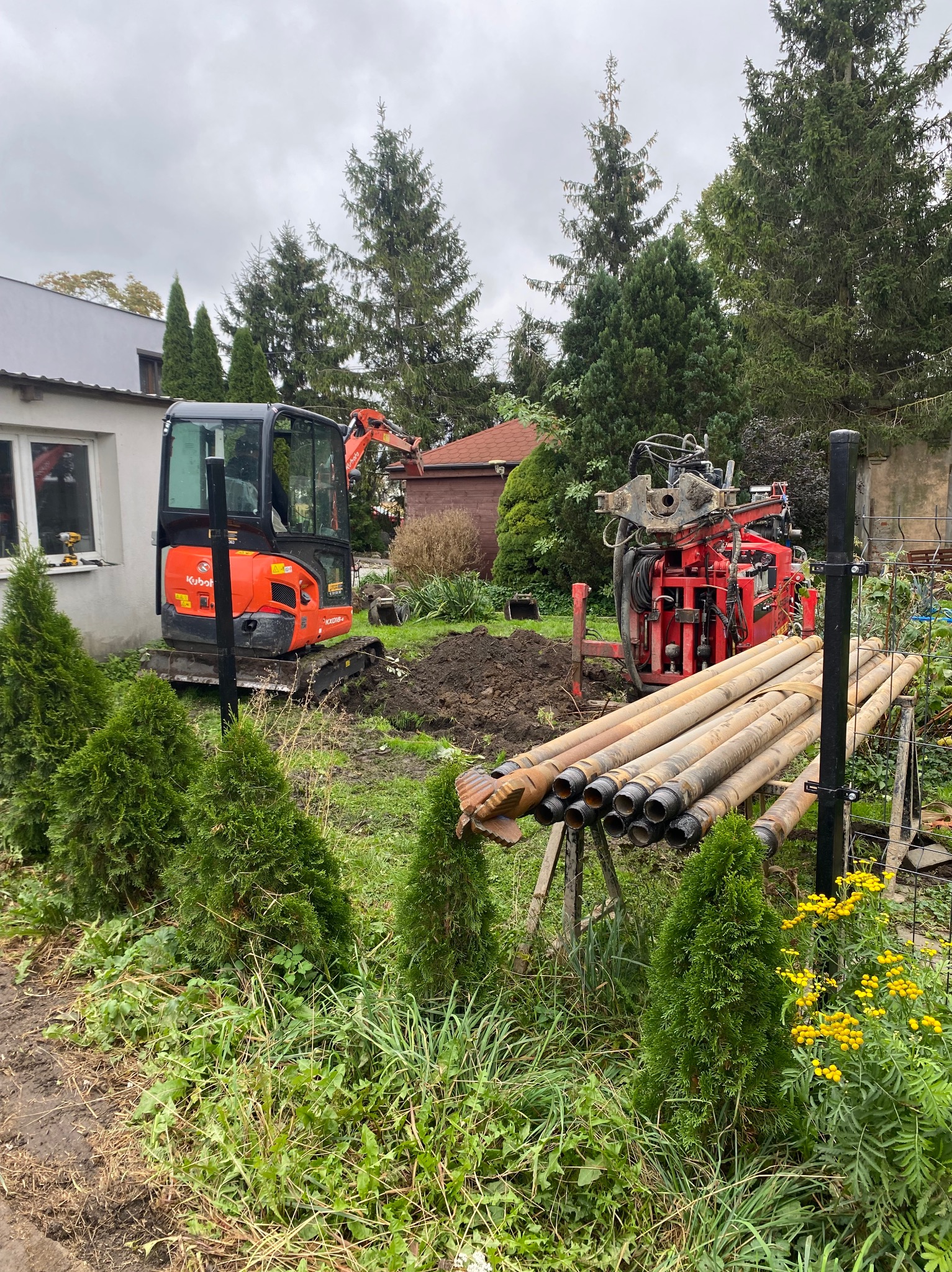 Czerwona wiertnica obok sterty rur wiertniczych na tle minikoparki Kubota i zieleni ogrodu, z widocznym wykopem w ziemi.