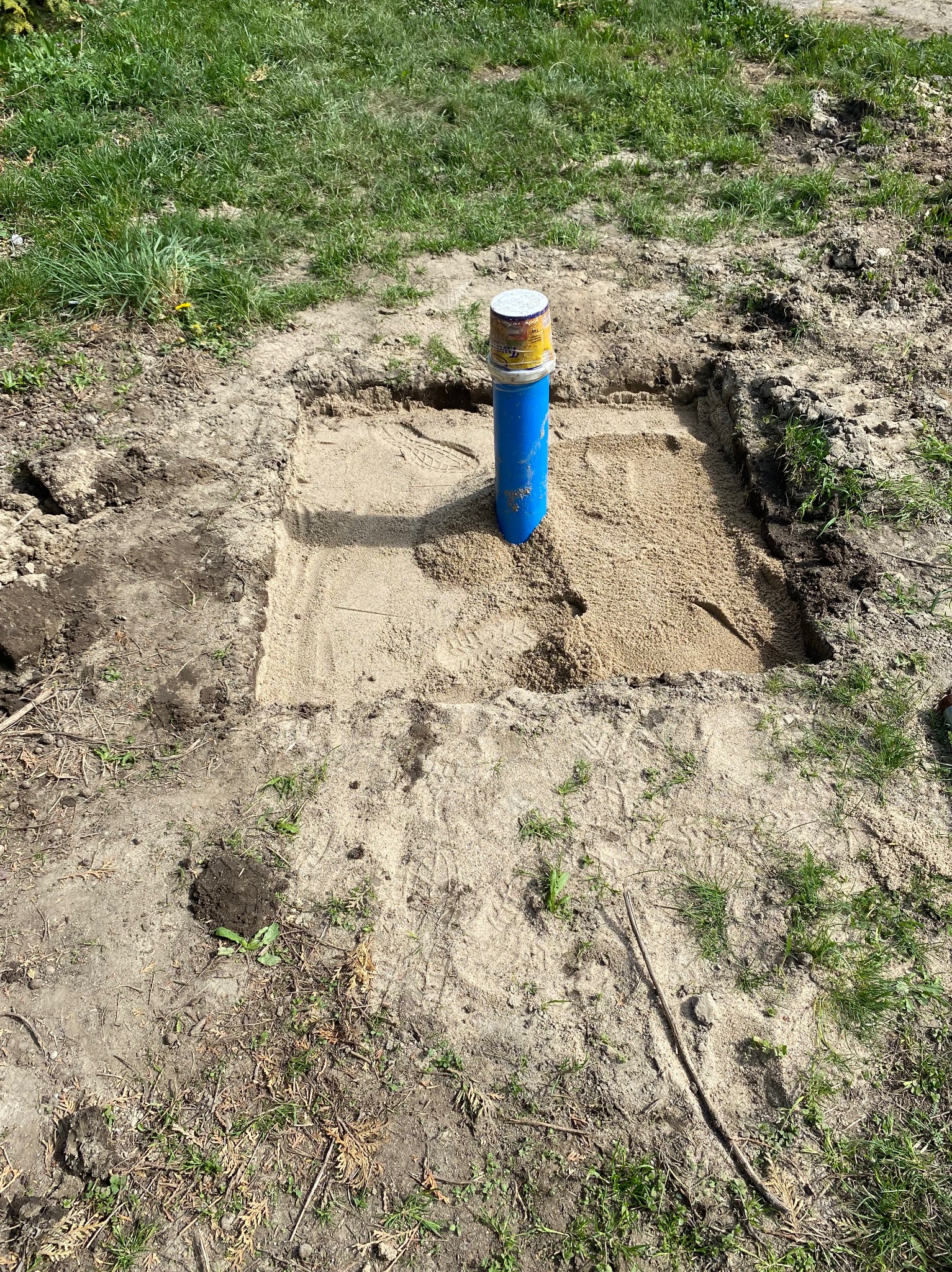 Niebieska rura wystaje z kwadratowego wykopu wypełnionego piaskiem na zewnątrz, otoczonego ziemią i trawą.