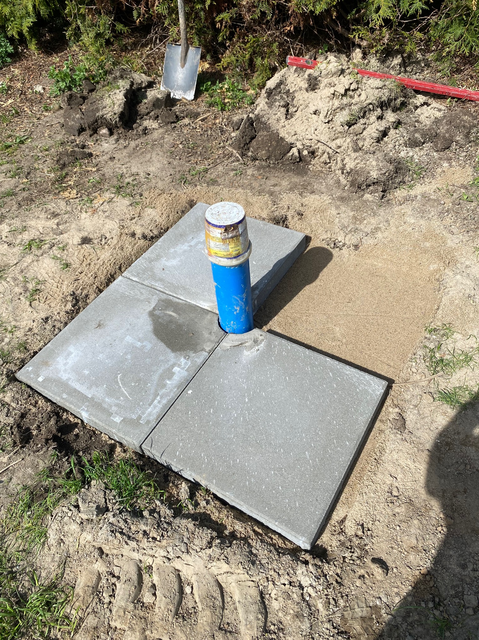 Nisko osadzona studnia z niebieską rurą wystającą ponad powierzchnię, otoczona czterema betonowymi płytami chodnikowymi, z widoczną łopatą i poziomicą w tle.