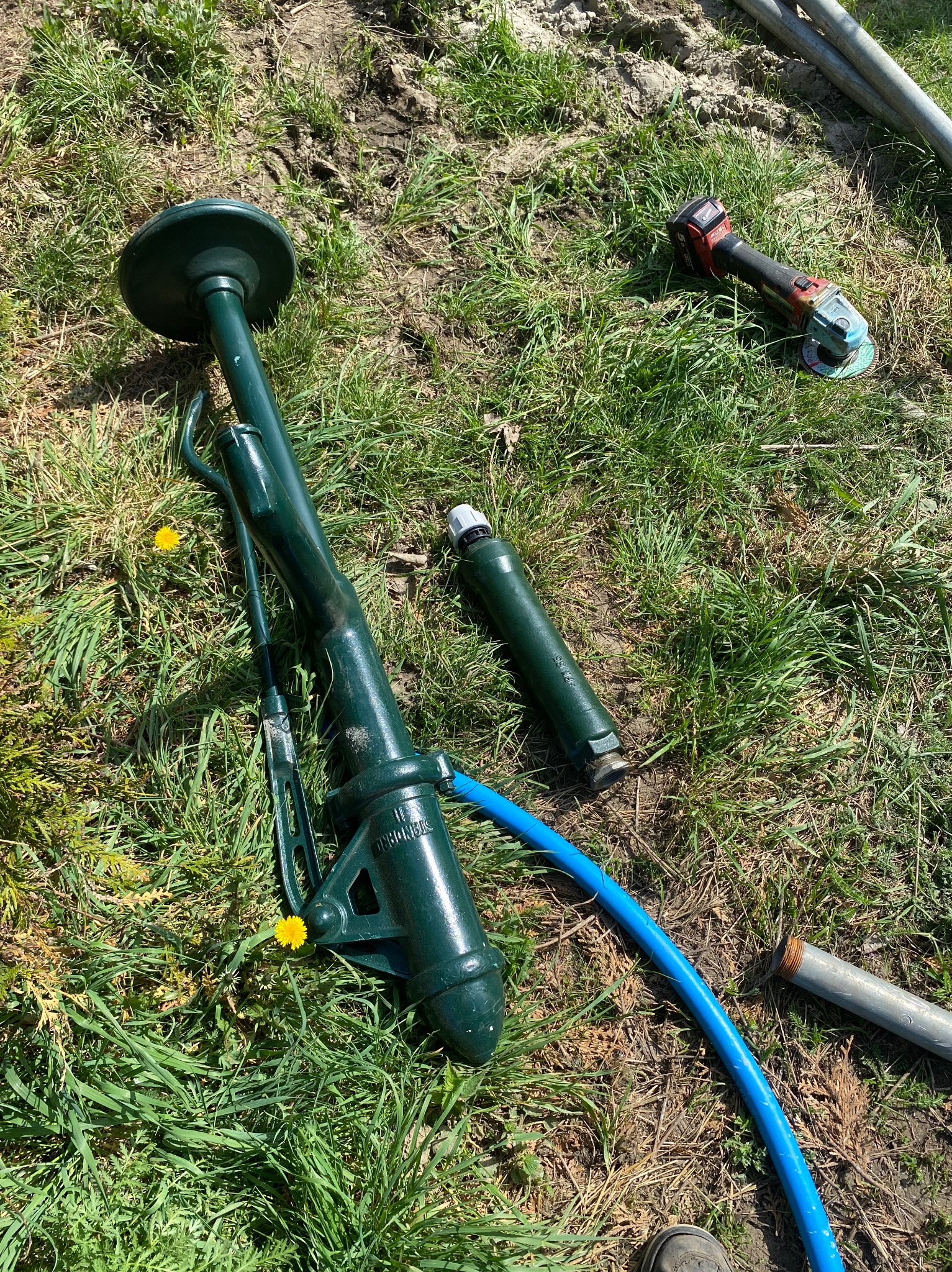 Elementy pompy abisyńskiej w kolorze ciemnej zieleni, rura, szlifierka kątowa i fragment buta na trawie w słoneczny dzień.