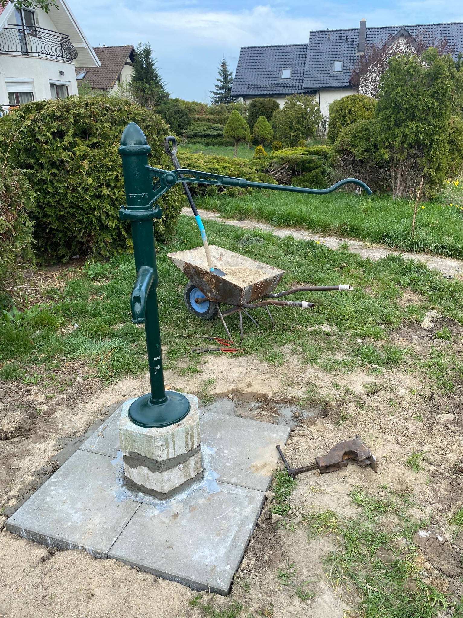 Zielona pompa ręczna zamontowana na betonowej podstawie, obok taczka z piaskiem i narzędzia, na tle domów jednorodzinnych i zieleni.
