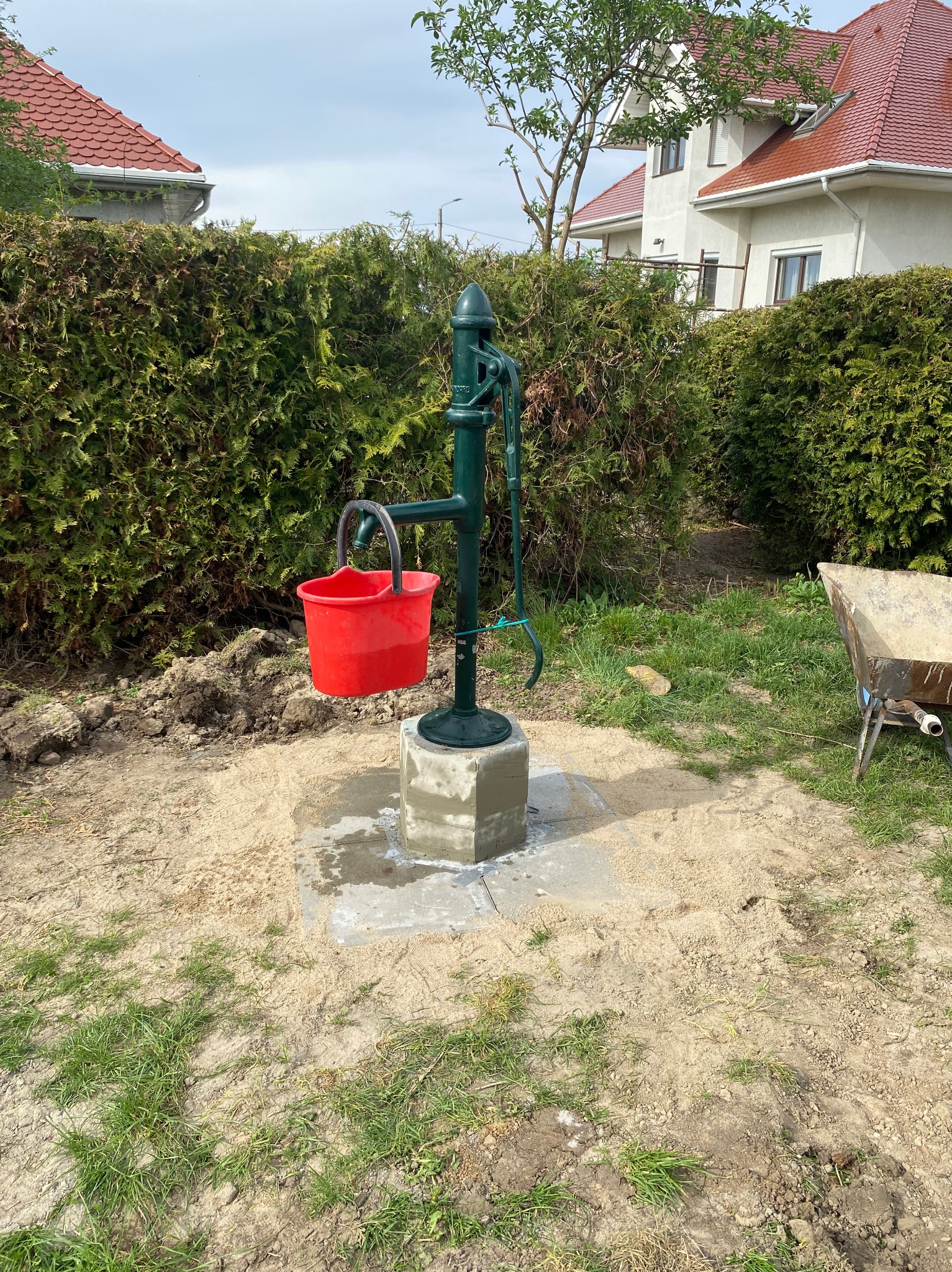 Zainstalowana zielona pompa ręczna z czerwonym wiadrem na betonowej podstawie, otoczona piaskiem i trawą, z żywopłotem w tle.