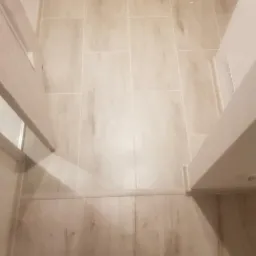 Jasne, prostokątne płytki podłogowe imitujące drewno, ułożone w korytarzu pomiędzy ościeżnicami drzwiowymi.