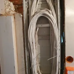 Instalacja elektryczna w trakcie montażu: białe kable ułożone w pętle wewnątrz metalowej skrzynki montażowej, widoczne czarne rury karbowane, ściana w stanie surowym z pianką montażową i otworem...