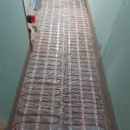 Rozłożona mata grzewcza z czerwonymi kablami na białej siatce w wąskim korytarzu, przygotowana do zalania wylewką.