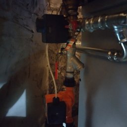 solar serwis dariusz borys - Fragment instalacji grzewczej z pompą obiegową i zaworami, zamontowany przy starej, nierównej ścianie z cegły i zaprawy.