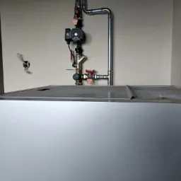 Szczeg&oacute;łowe ujęcie instalacji grzewczej z rurami i zaworami, zamontowanej nad metalową obudową pieca, na tle jasnej ściany. Widoczne elementy łączeniowe i przewody.