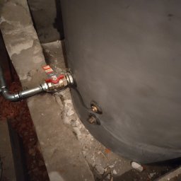 solar serwis dariusz borys - Szary zbiornik z podłączonym zaworem odcinającym, widoczne fragmenty instalacji w surowym otoczeniu. Detal montażu w piwnicy.