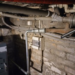 solar serwis dariusz borys - Instalacja wodna z nową, pionową rurą doprowadzoną do zaworu i skrzynki elektrycznej na tle starej, ceglanej ściany. Widoczne inne rury i kable.