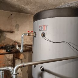 solar serwis dariusz borys - Szary bojler OKHT z podłączonymi rurami i zaworami w piwnicy z surowym betonowym sufitem i ścianami. Widoczne ślady wilgoci.