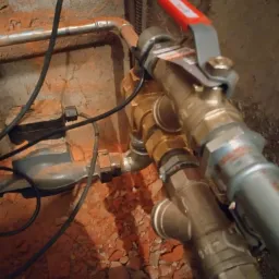 Złożony układ rur z zaworem z czerwoną rączką w surowym wnętrzu. Widoczne elementy instalacji, przewody i fragmenty gruzu na podłożu. Detal instalacji hydraulicznej.