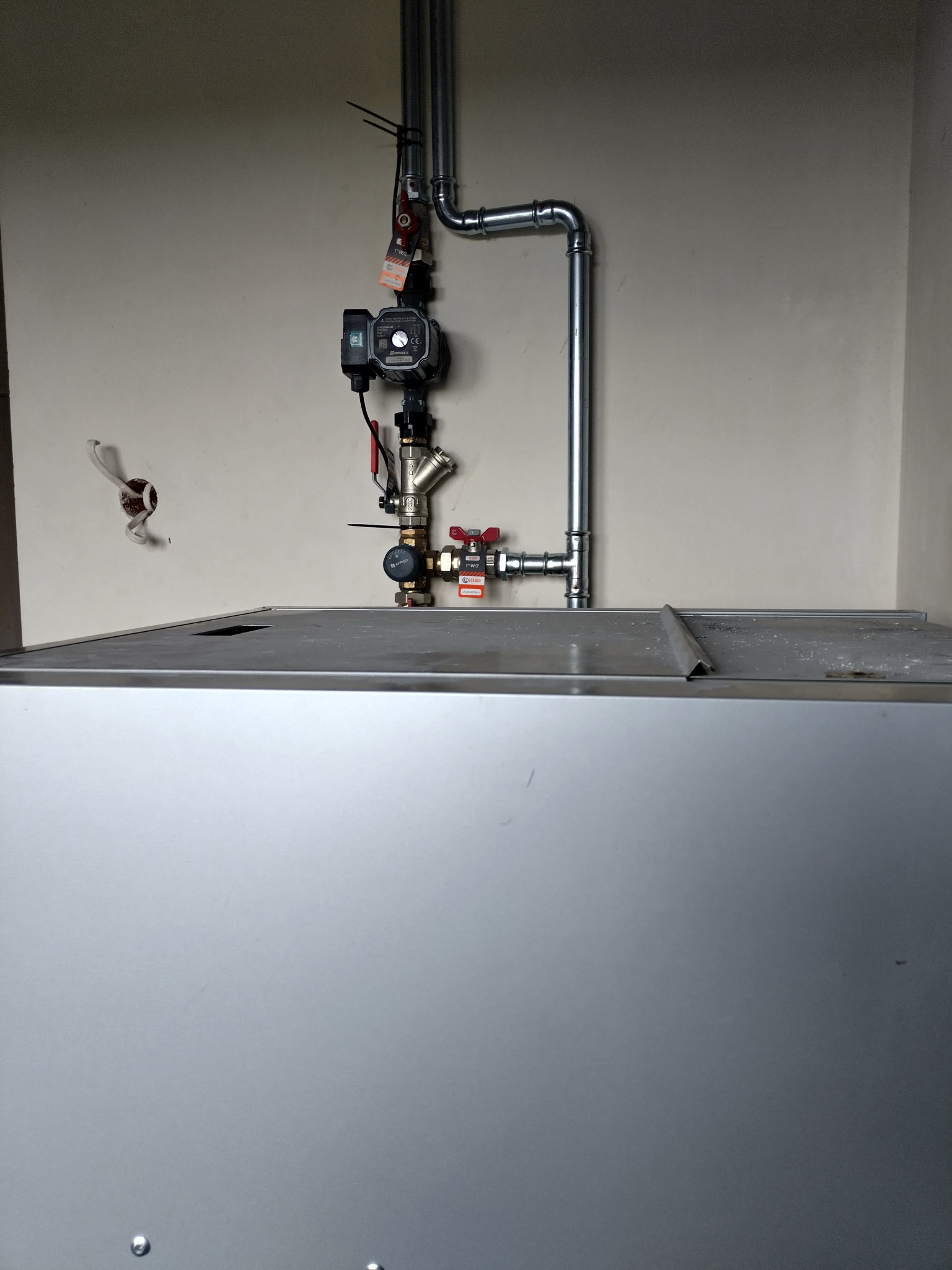 Szczegółowe ujęcie instalacji grzewczej z rurami i zaworami, zamontowanej nad metalową obudową pieca, na tle jasnej ściany. Widoczne elementy łączeniowe i przewody.