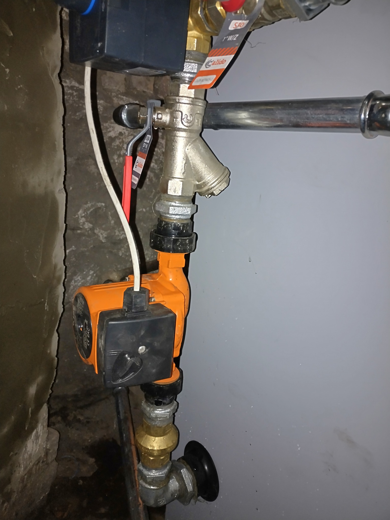 Pompa obiegowa Calido zamontowana na rurach instalacji grzewczej, widoczne zawory i przewody. Elementy instalacji na tle ściany pomalowanej na szaro.