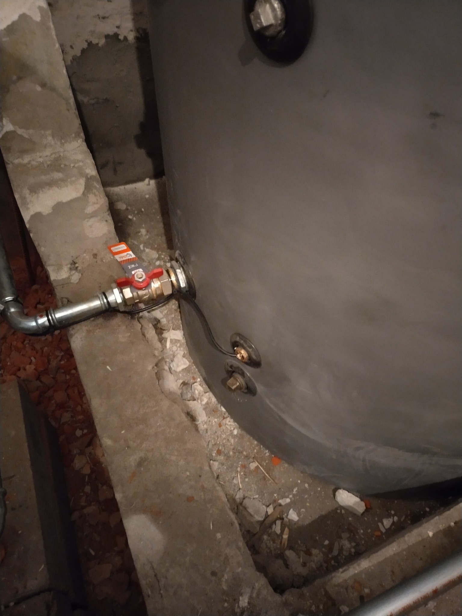 Szary zbiornik z podłączonym zaworem odcinającym, widoczne fragmenty instalacji w surowym otoczeniu. Detal montażu w piwnicy.