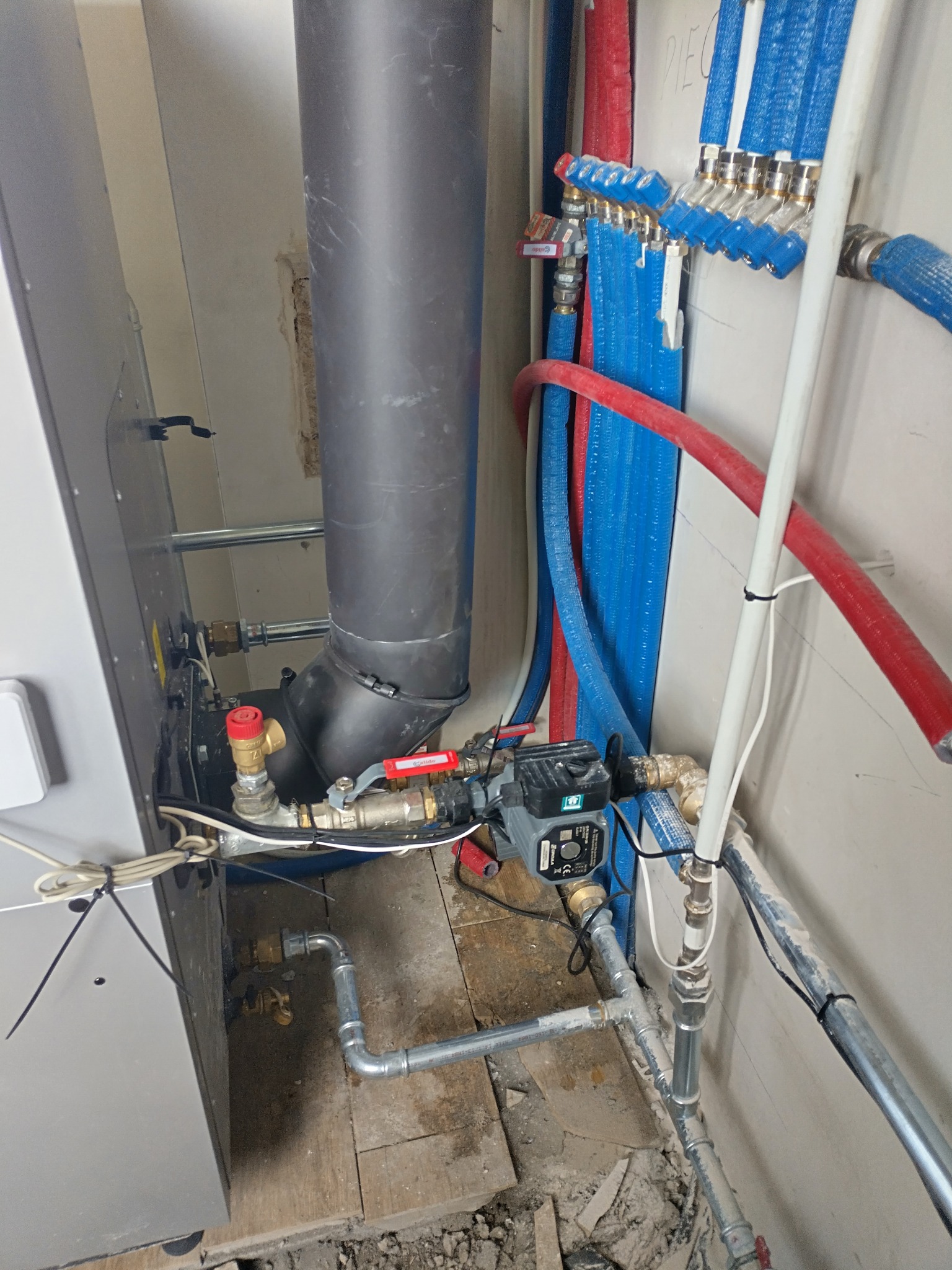 Szczegółowe ujęcie instalacji grzewczej z piecem, pompą obiegową i rurami w kolorach niebieskim i czerwonym, ukazujące złożoność systemu w pomieszczeniu technicznym.