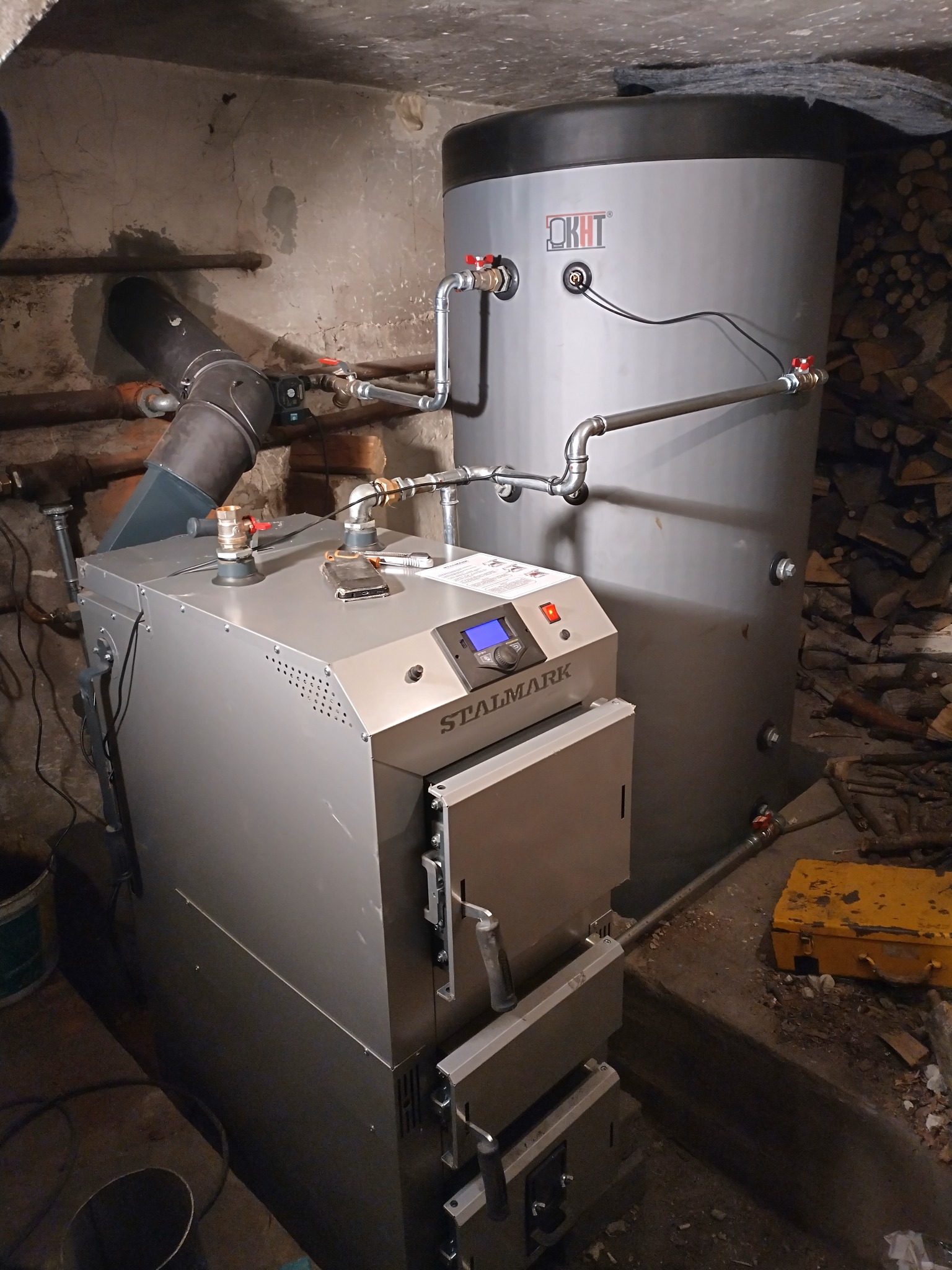 Szary piec CO marki Stalmark z zasobnikiem OKHT, połączony rurami. Obok leżą kawałki drewna i żółta skrzynka narzędziowa. Ściany i sufit w surowym stanie.