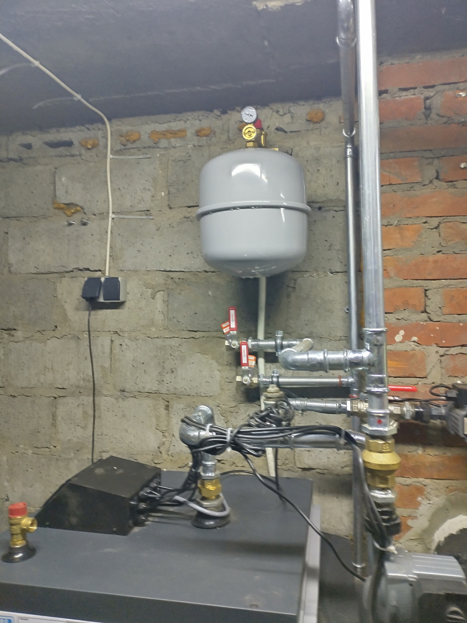 Zamontowany zbiornik wyrównawczy z manometrem i instalacją hydrauliczną na tle ściany z cegieł i betonu komórkowego w piwnicy.