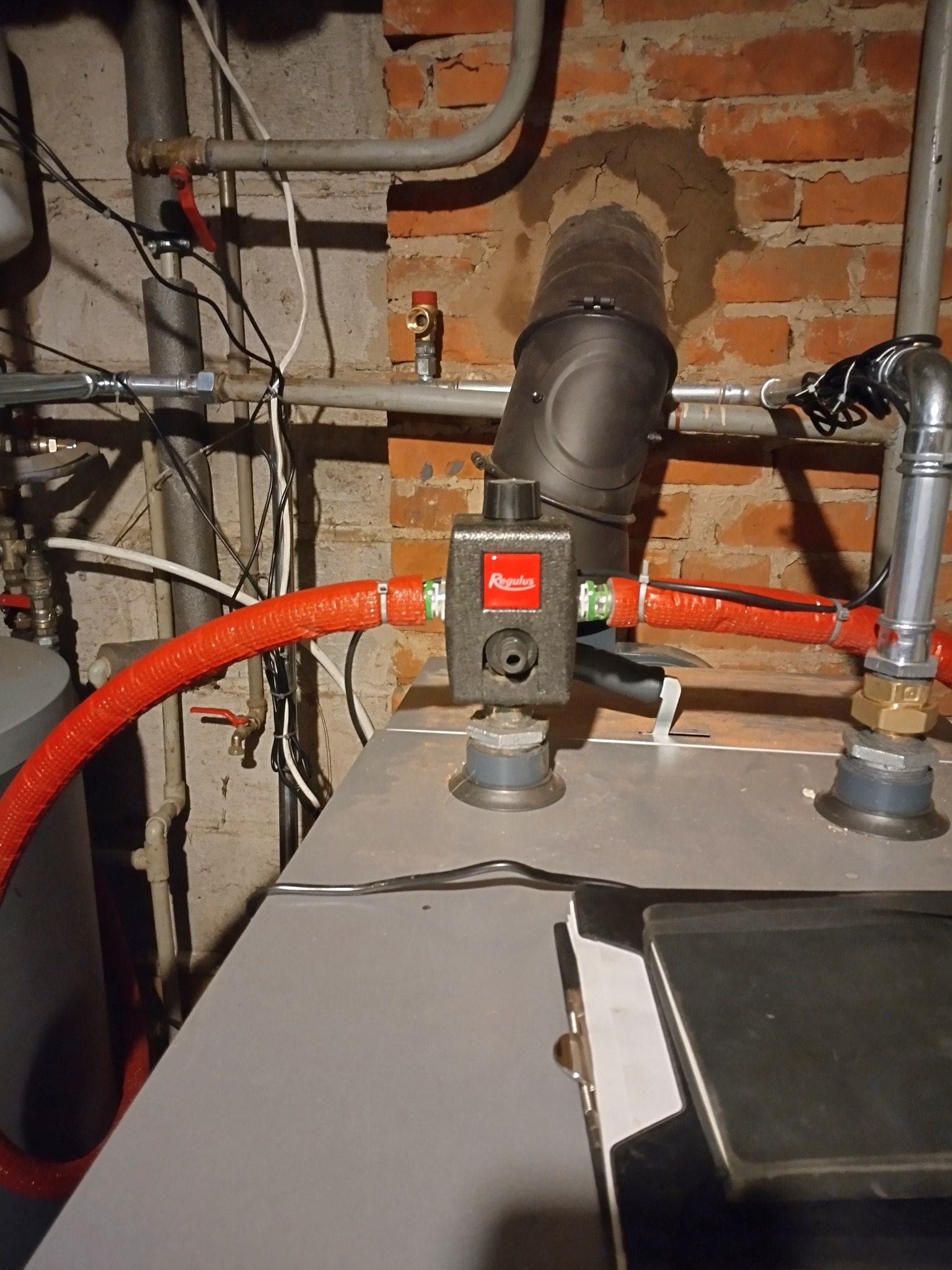 Zamontowany zawór Regulus na kotle grzewczym z czerwonymi rurami w piwnicy z ceglaną ścianą. Widoczne elementy instalacji i rury.
