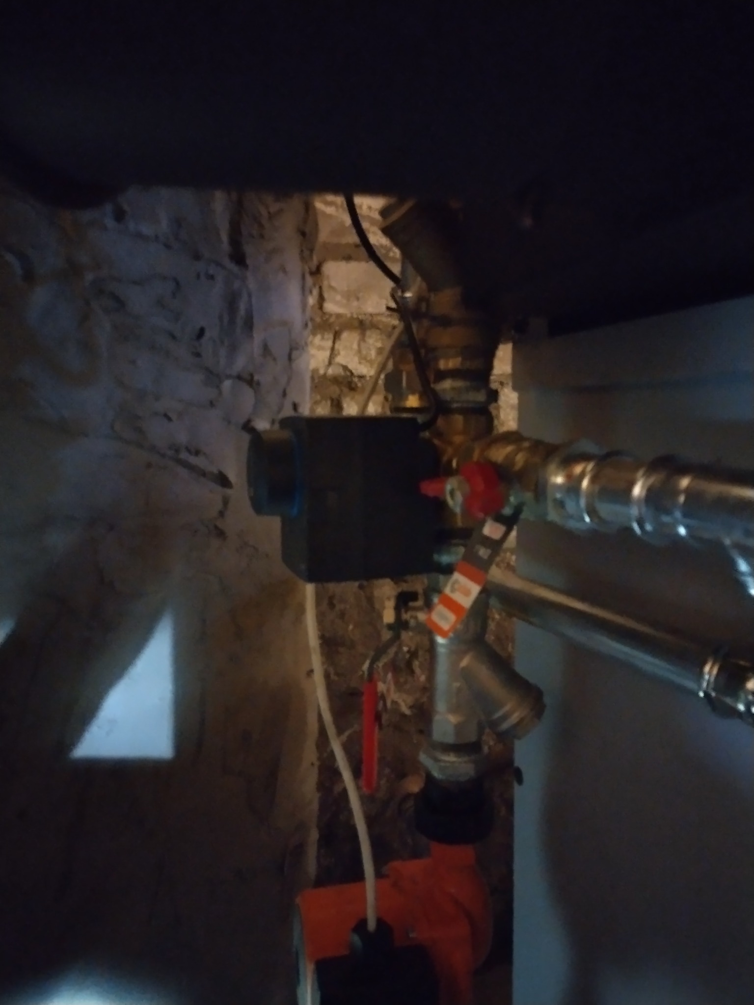 Fragment instalacji grzewczej z zaworami i czarną skrzynką sterującą, w pomieszczeniu z surową ścianą z cegły i słabym oświetleniem.