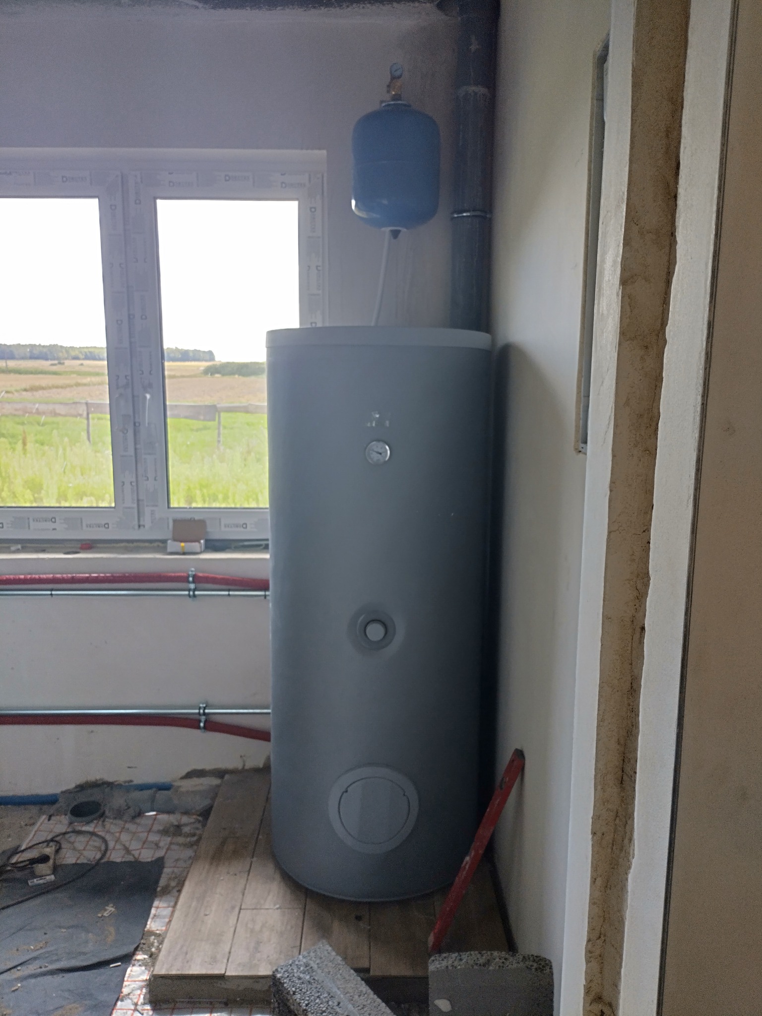 Szary bojler z niebieskim naczyniem wyrównawczym obok okna, widoczne rury instalacji CO i fragment ogrzewania podłogowego na podłodze w surowym wnętrzu.