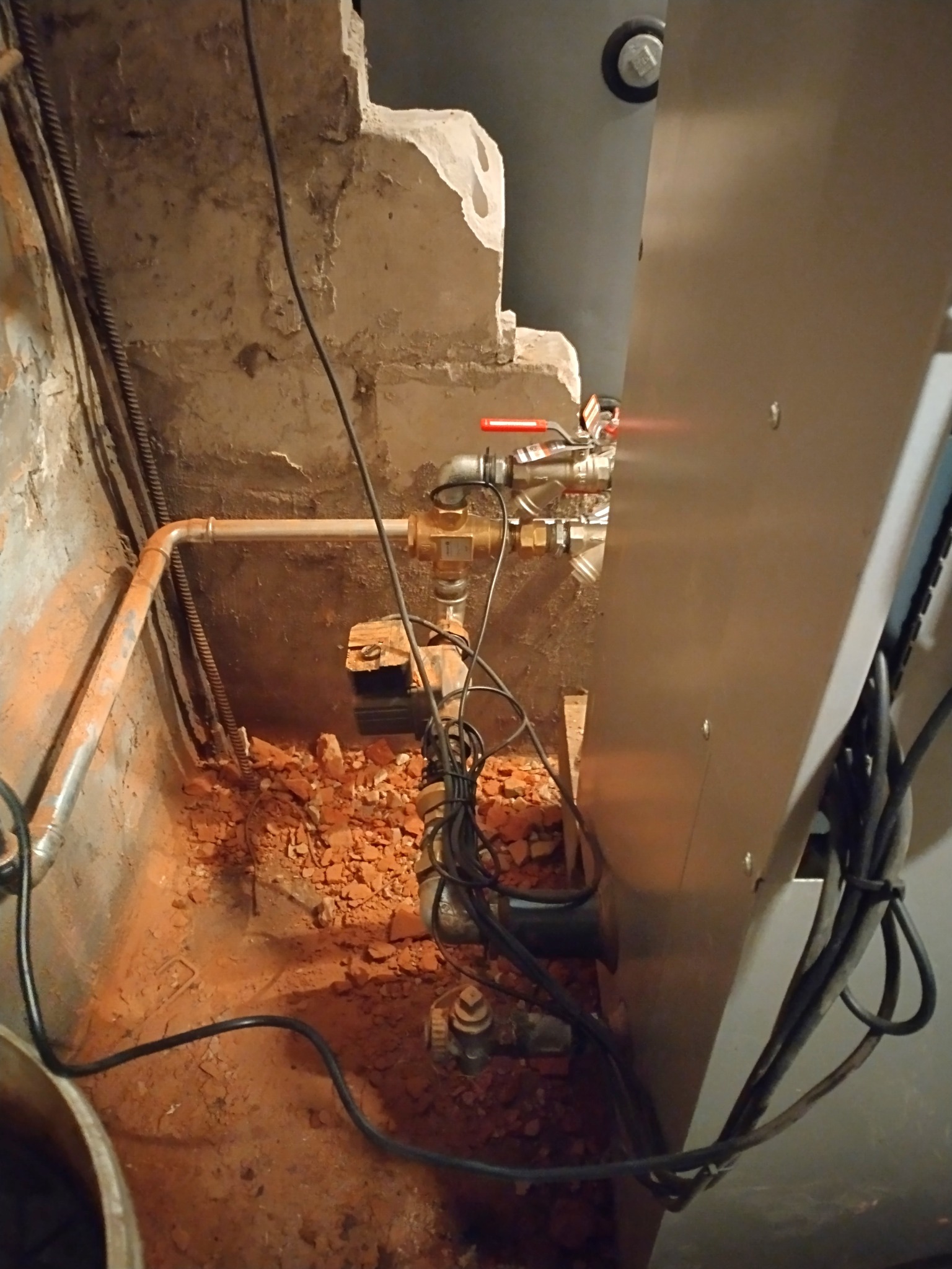 Fragment instalacji CO z zaworami, rurami miedzianymi, przewodami elektrycznymi i gruzem na podłodze w pomieszczeniu technicznym.