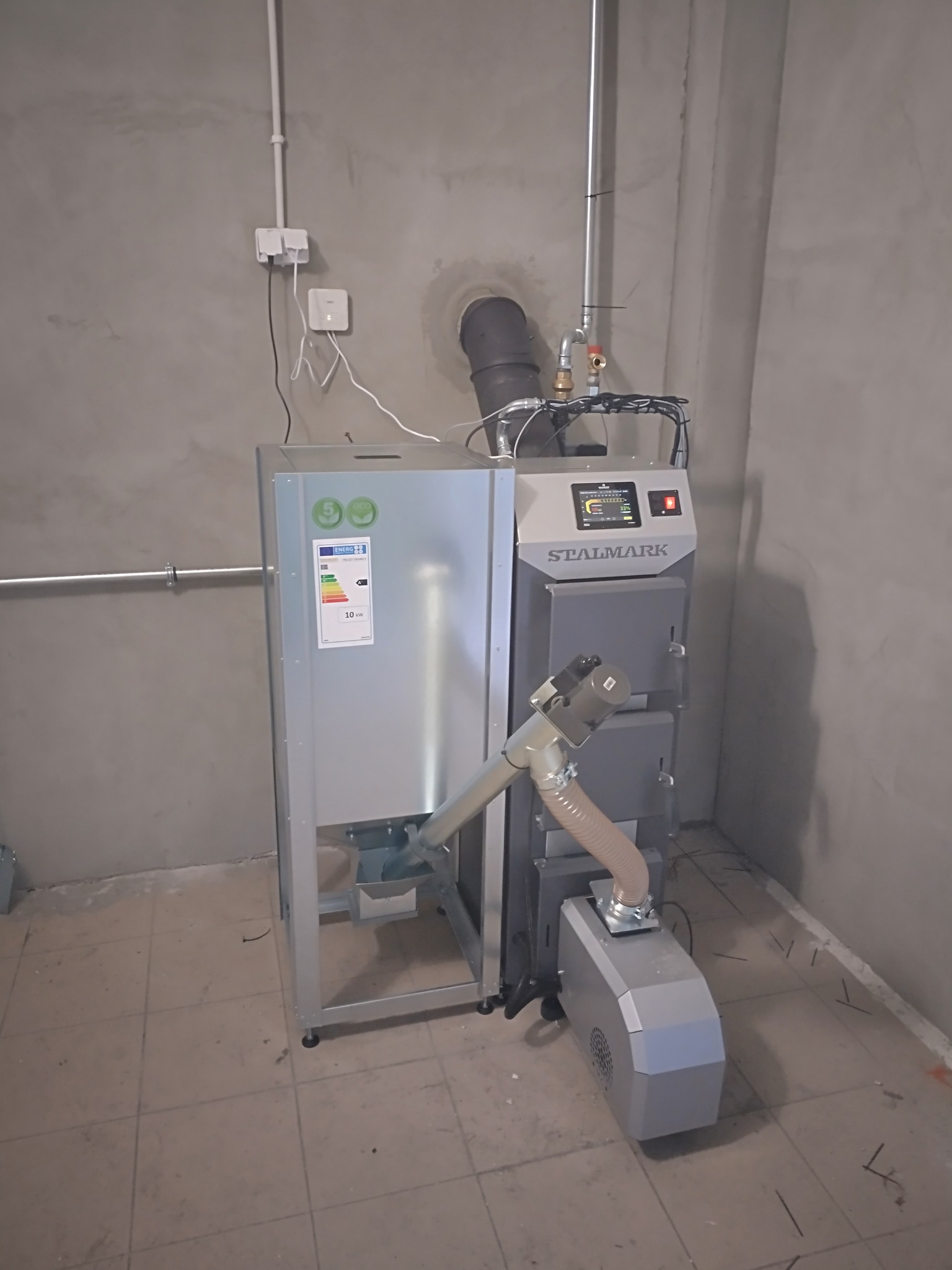 Stalmark - piec na pellet z podajnikiem, zainstalowany w pomieszczeniu z szarą, surową ścianą i brązową podłogą z płytek. Widoczna etykieta energetyczna.
