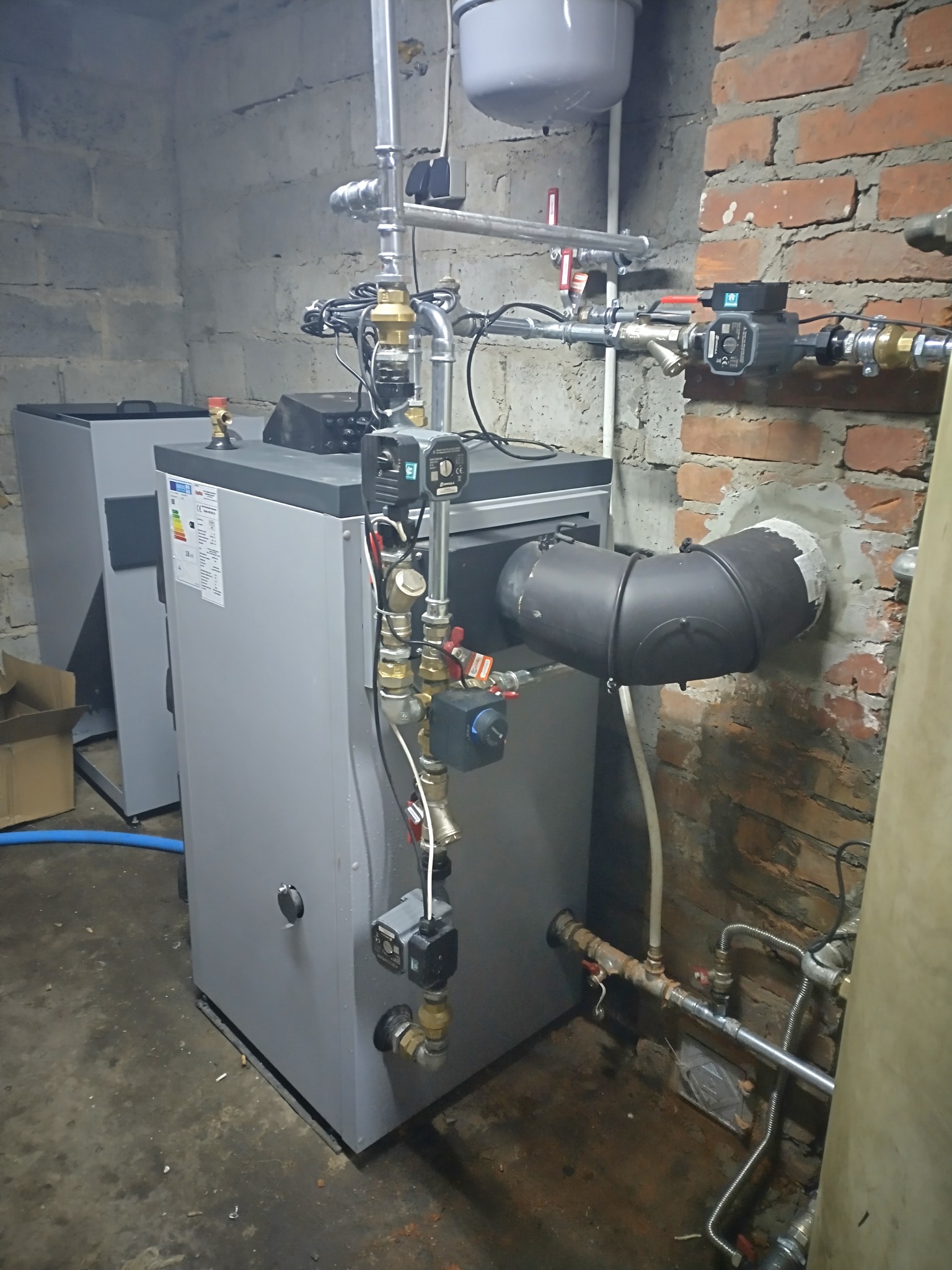 Szary piec CO z podłączeniami w kotłowni w Krasnystawie. Widoczne rury, zawory i elementy sterujące. Ściany z cegły i betonu. Ukończona instalacja grzewcza.