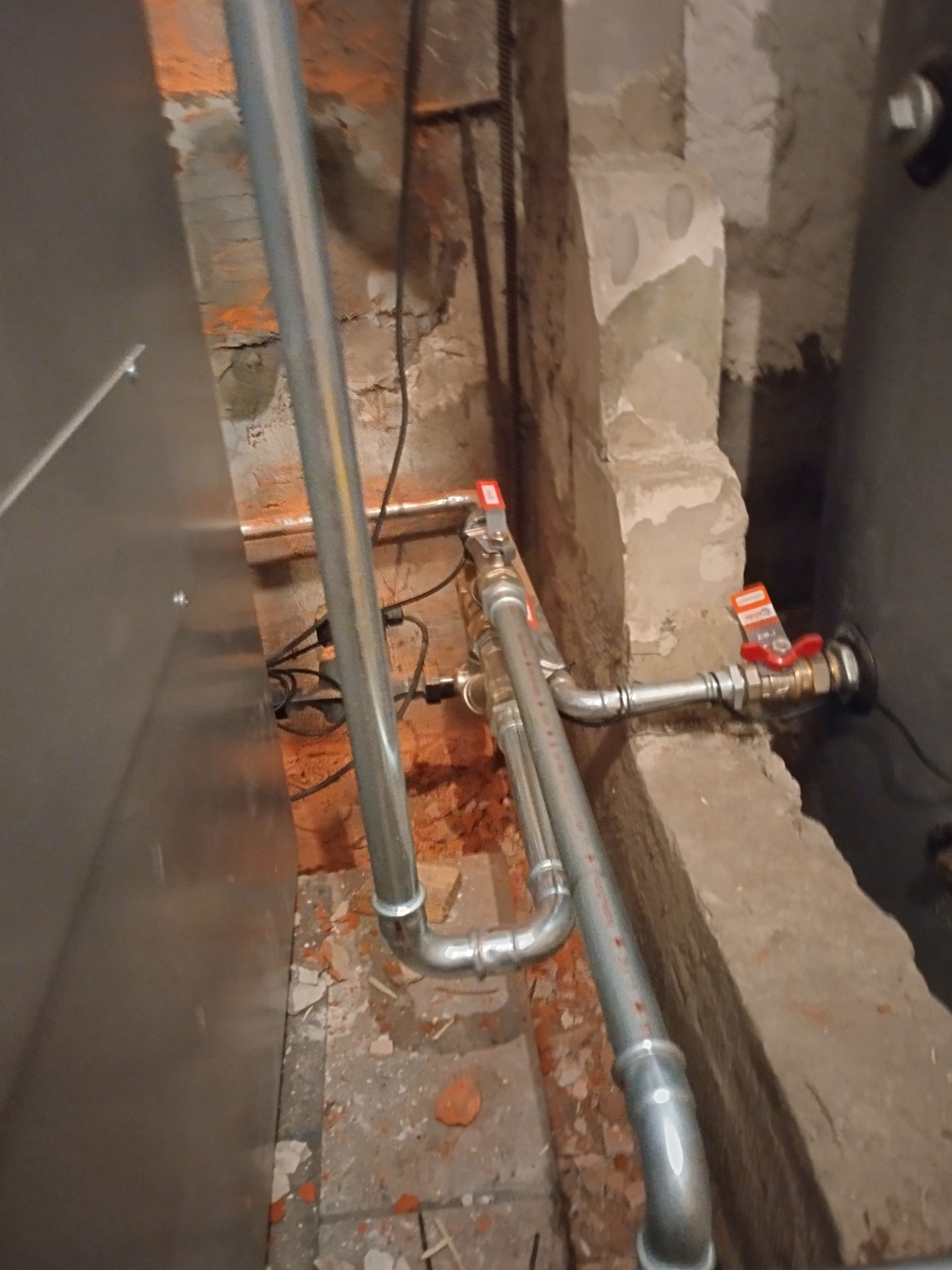 Szare rury instalacji CO z zaworami, prowadzone wzdłuż ściany z cegły i betonu. Fragment pieca widoczny po lewej, instalacja po prawej stronie.