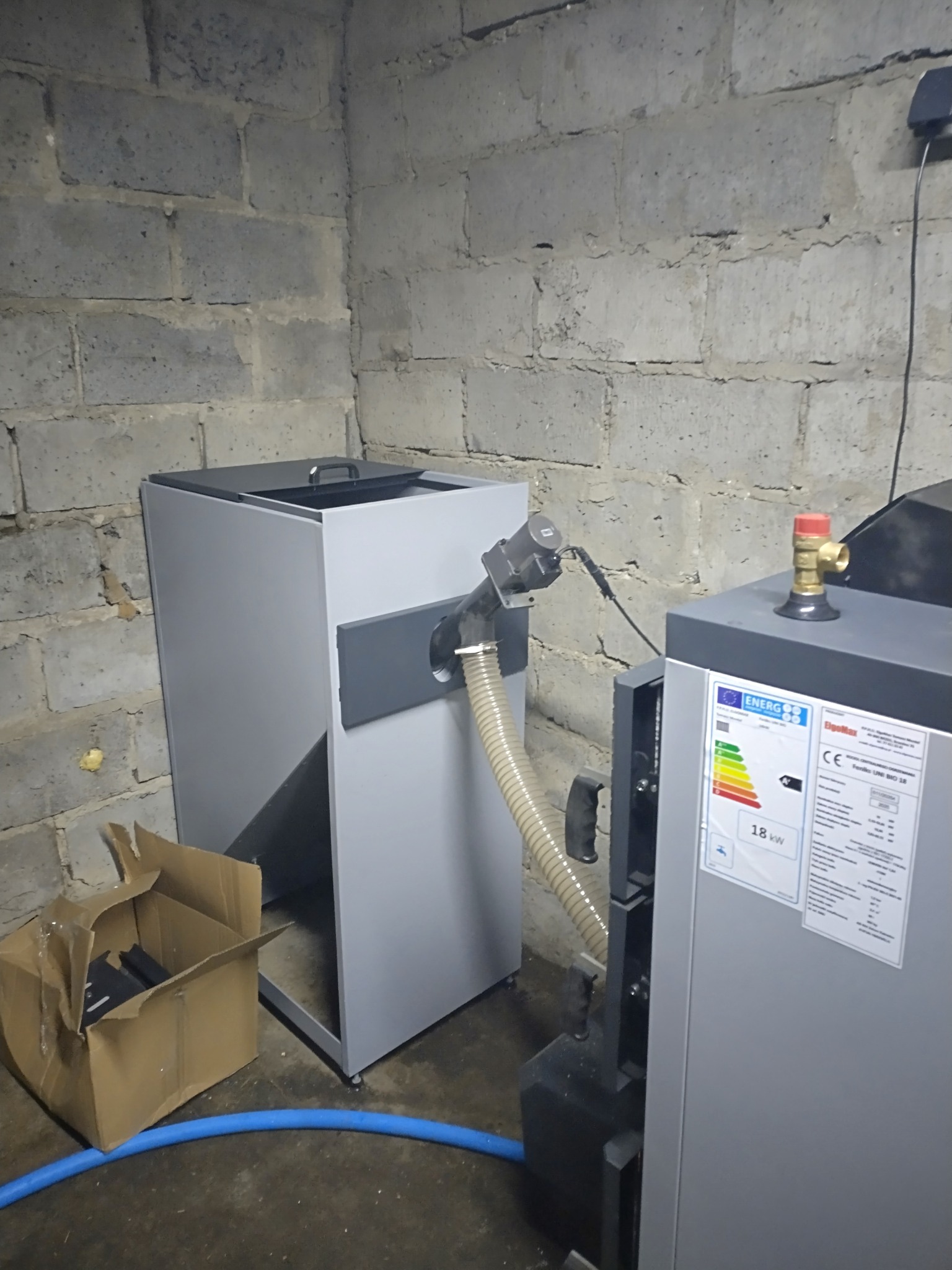 Szary piec na paliwo stałe z podajnikiem, ustawiony w pomieszczeniu z betonowych pustaków. Widoczna etykieta energetyczna 18kW i karton obok.
