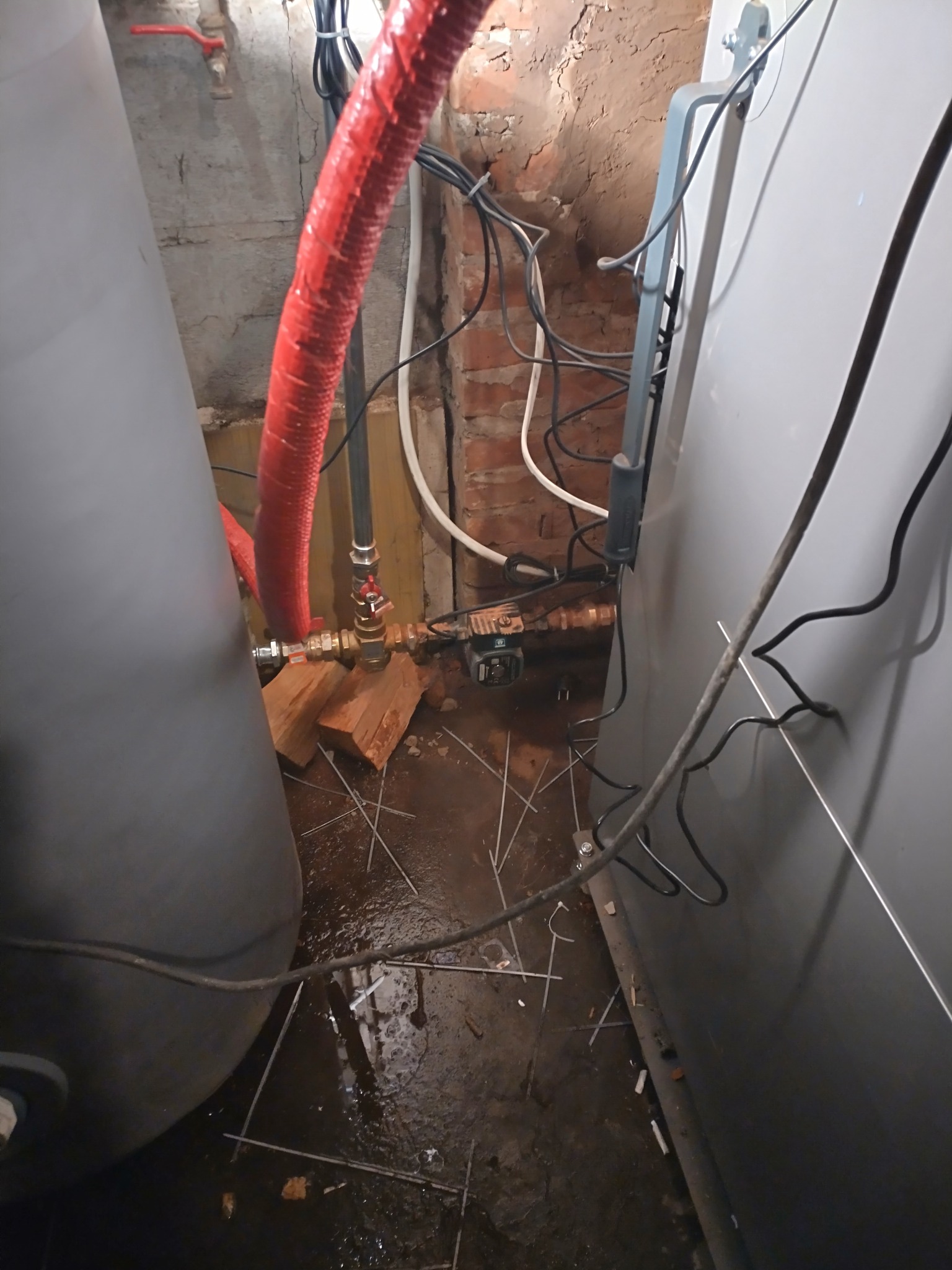 Szara obudowa pieca CO, czerwona rura izolacyjna, fragment instalacji hydraulicznej na tle ceglanej ściany. Widoczne przewody elektryczne i elementy montażowe.