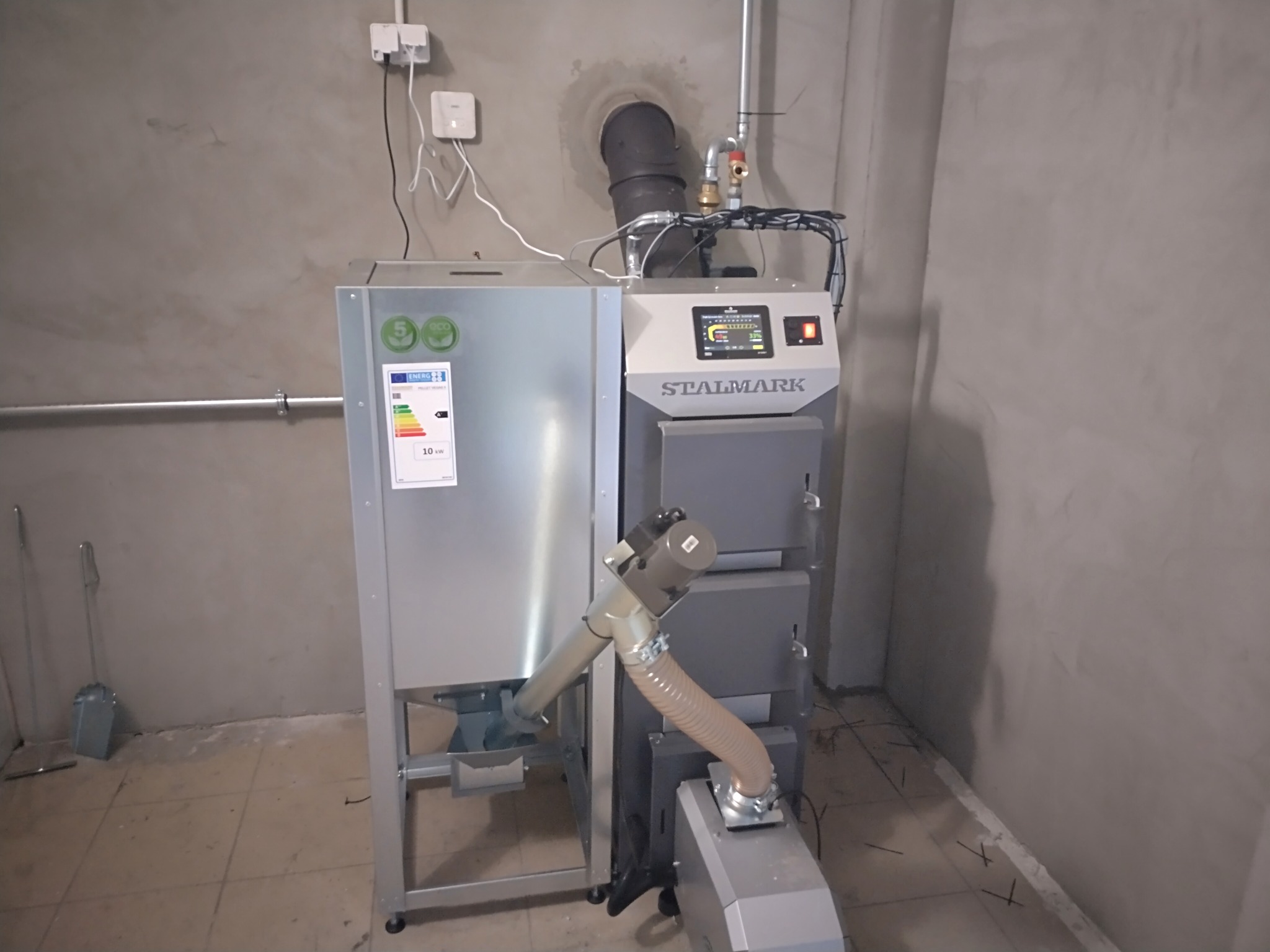 Stalmark: Nowoczesny piec CO na ekogroszek z podajnikiem ślimakowym, zainstalowany w surowym pomieszczeniu z betonową ścianą i widocznymi przewodami.