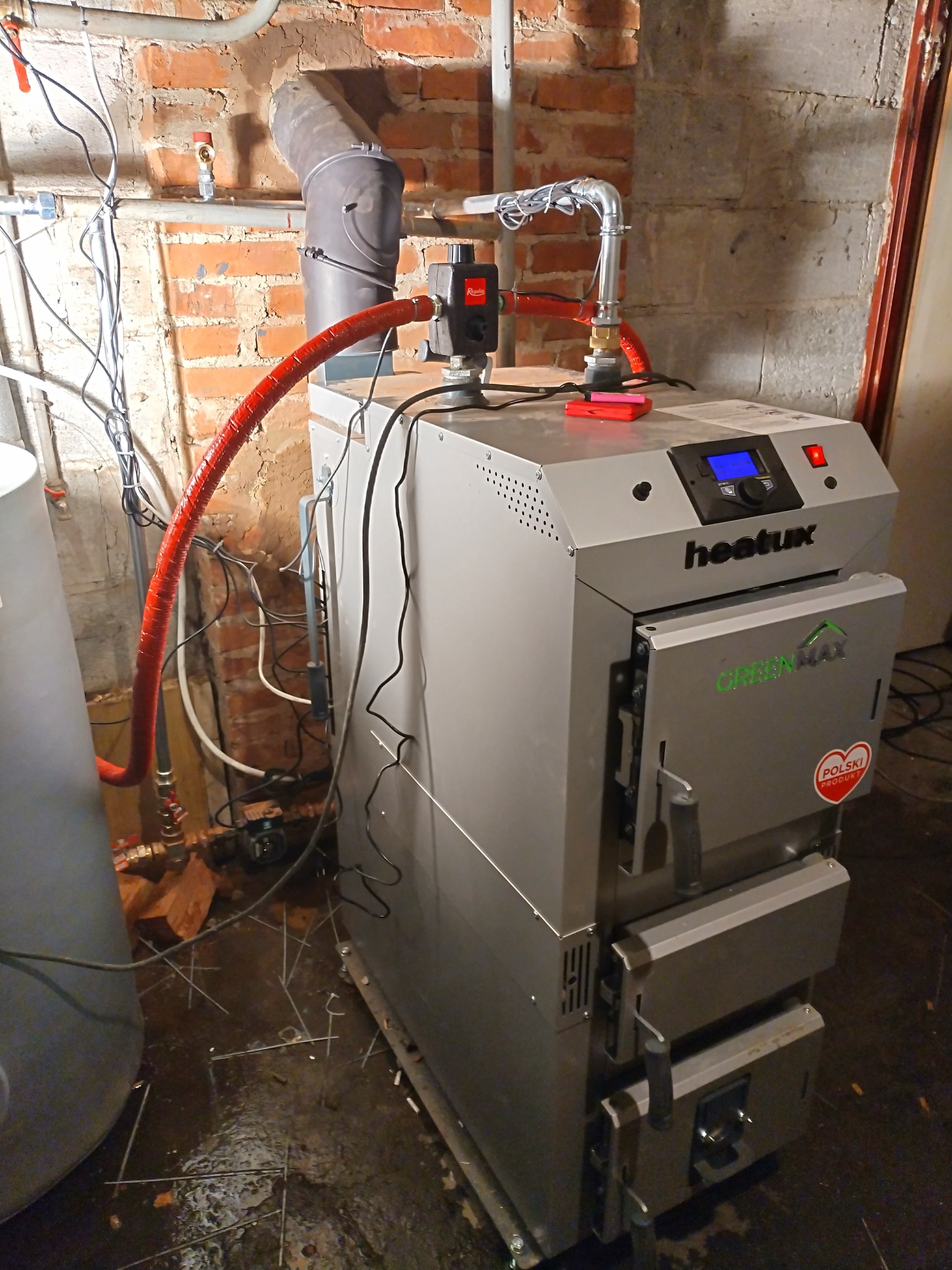 Szary piec CO marki Heatux Green Max z czerwoną izolacją rury w kotłowni z ceglaną ścianą i wilgotną posadzką. Widoczne elementy instalacji.