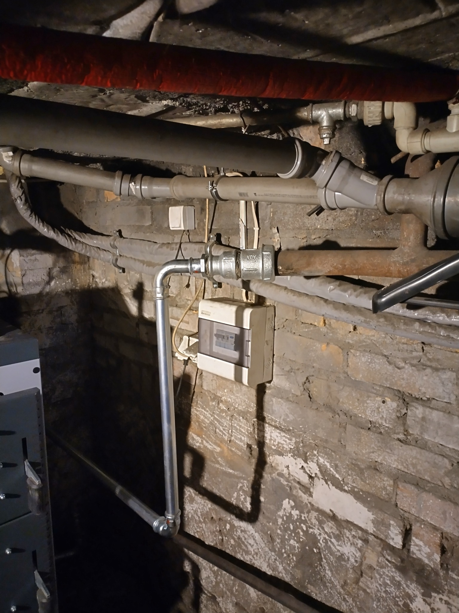 Instalacja wodna z nową, pionową rurą doprowadzoną do zaworu i skrzynki elektrycznej na tle starej, ceglanej ściany. Widoczne inne rury i kable.