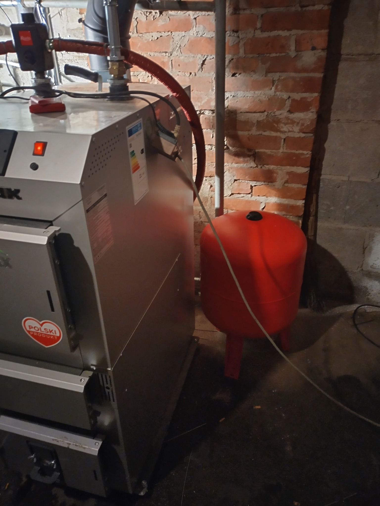 Nowoczesny piec CO z czerwonym zbiornikiem wyrównawczym na tle ściany z cegły. Widoczny fragment instalacji i oznaczenie 'Polski Produkt'.