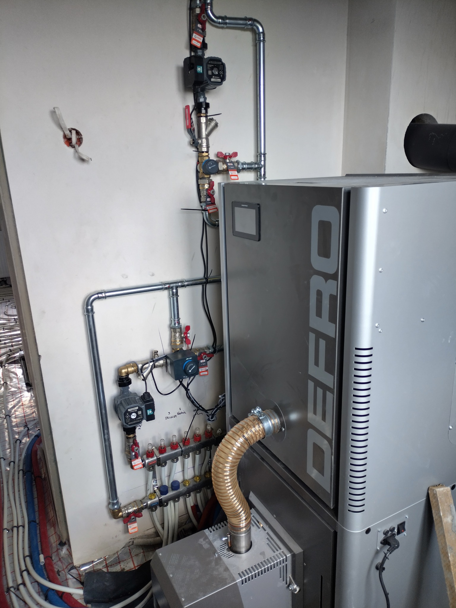 Szary piec Defro z podłączeniami, rurami, zaworami i elementami instalacji grzewczej. Widoczne ogrzewanie podłogowe w trakcie montażu.