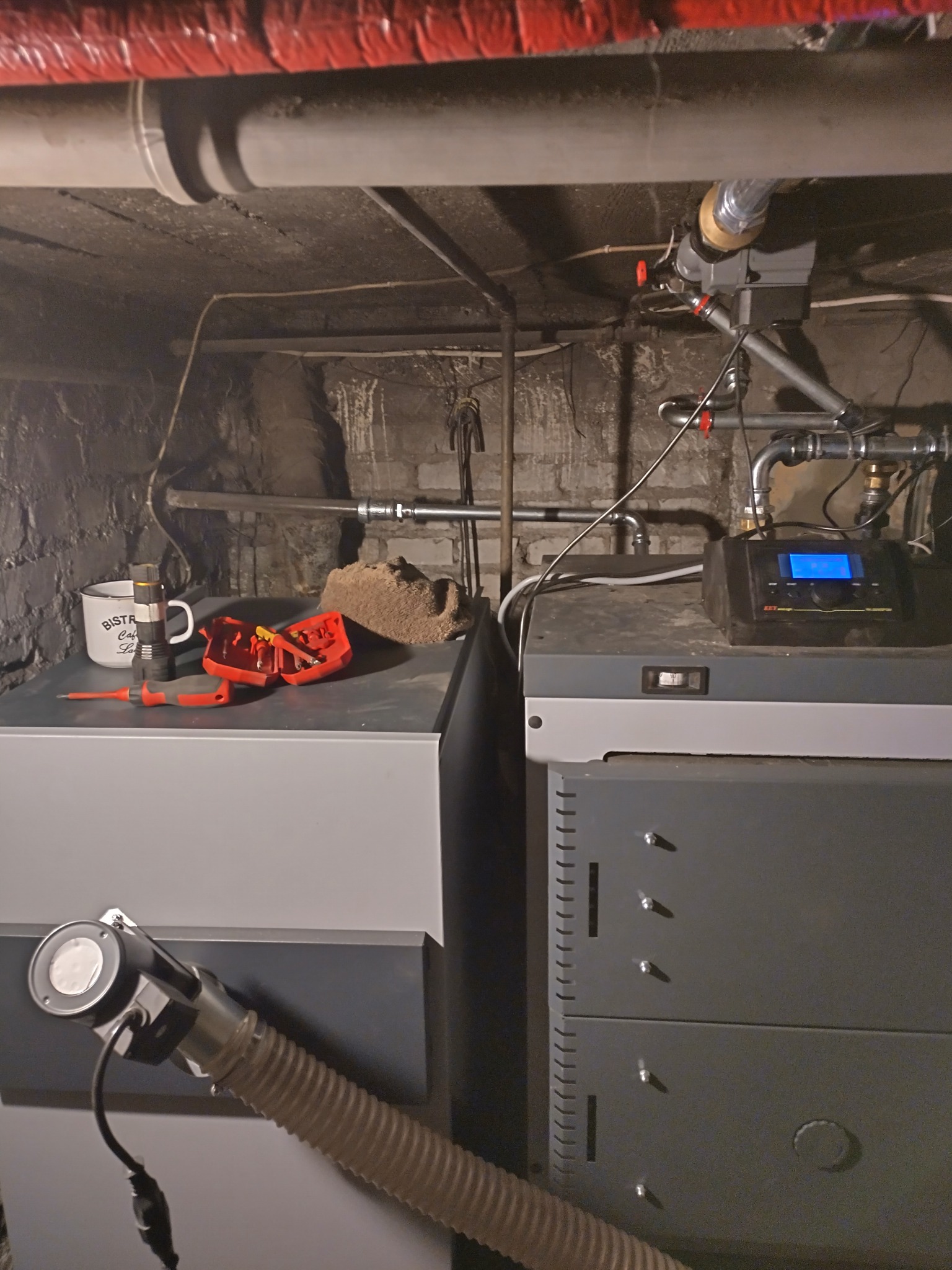 Dwa kotły grzewcze w piwnicy, na jednym narzędzia i kubek, rury instalacji grzewczej pod sufitem. Widoczne elementy instalacji i kotłowni.