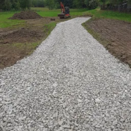 Świeżo ułożona, szara, kamienista droga dojazdowa na tle zielonego pola i lasu, z widocznym małym koparką w oddali.