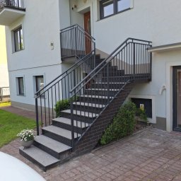 „Żelazny” Uslugi Spawalnicze - Zewnętrzne schody z szarego kamienia z czarną, metalową balustradą prowadzące do wejścia budynku. U podnóża schodów donica z białymi kwiatami.