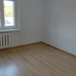 Pusty pokój po remoncie: panele podłogowe w odcieniach beżu, białe ściany, grzejnik żeberkowy pod oknem z niebieską roletą.