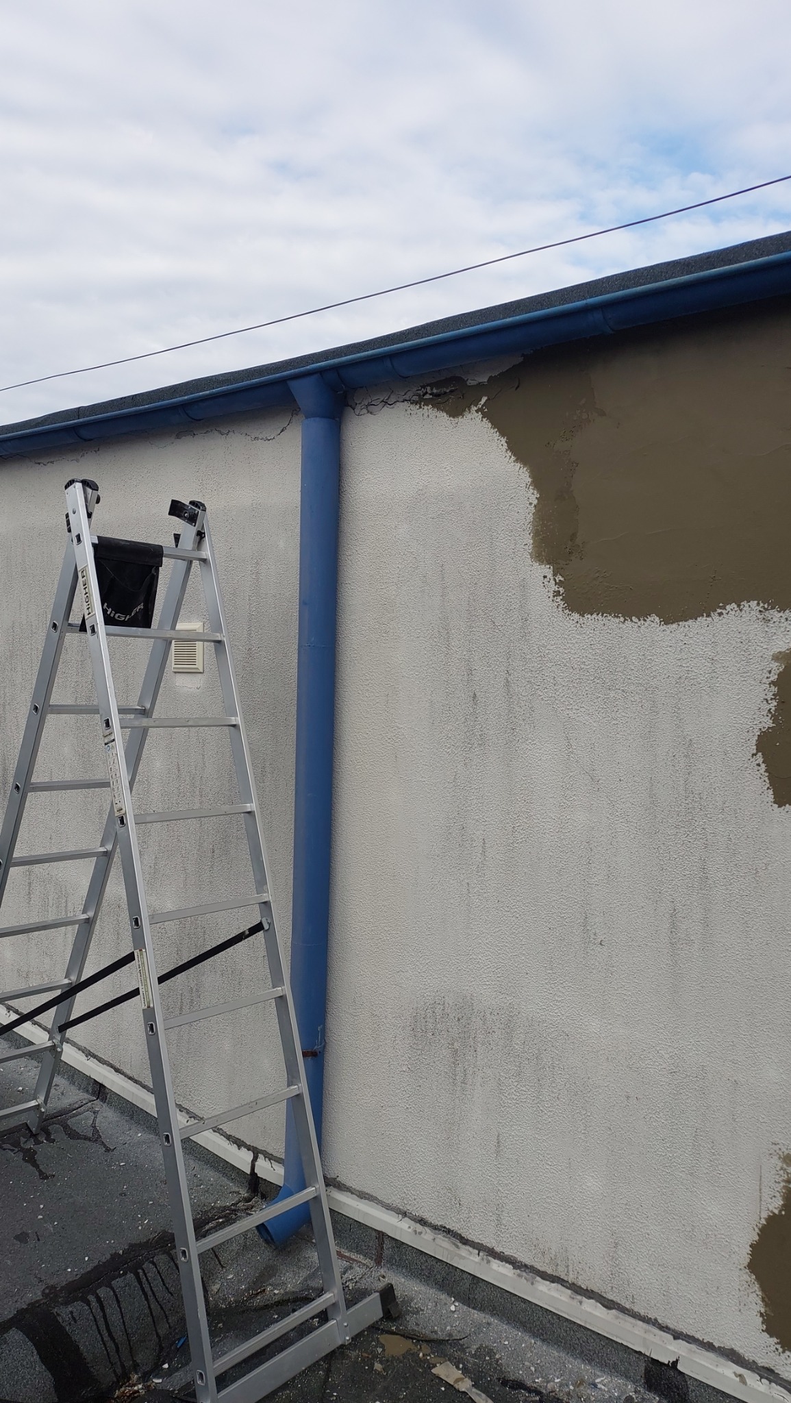 Ściana budynku z nałożonymi plamami szpachli, aluminiowa drabina przystawiona do ściany, niebieska rynna spustowa, fragment dachu z papy.