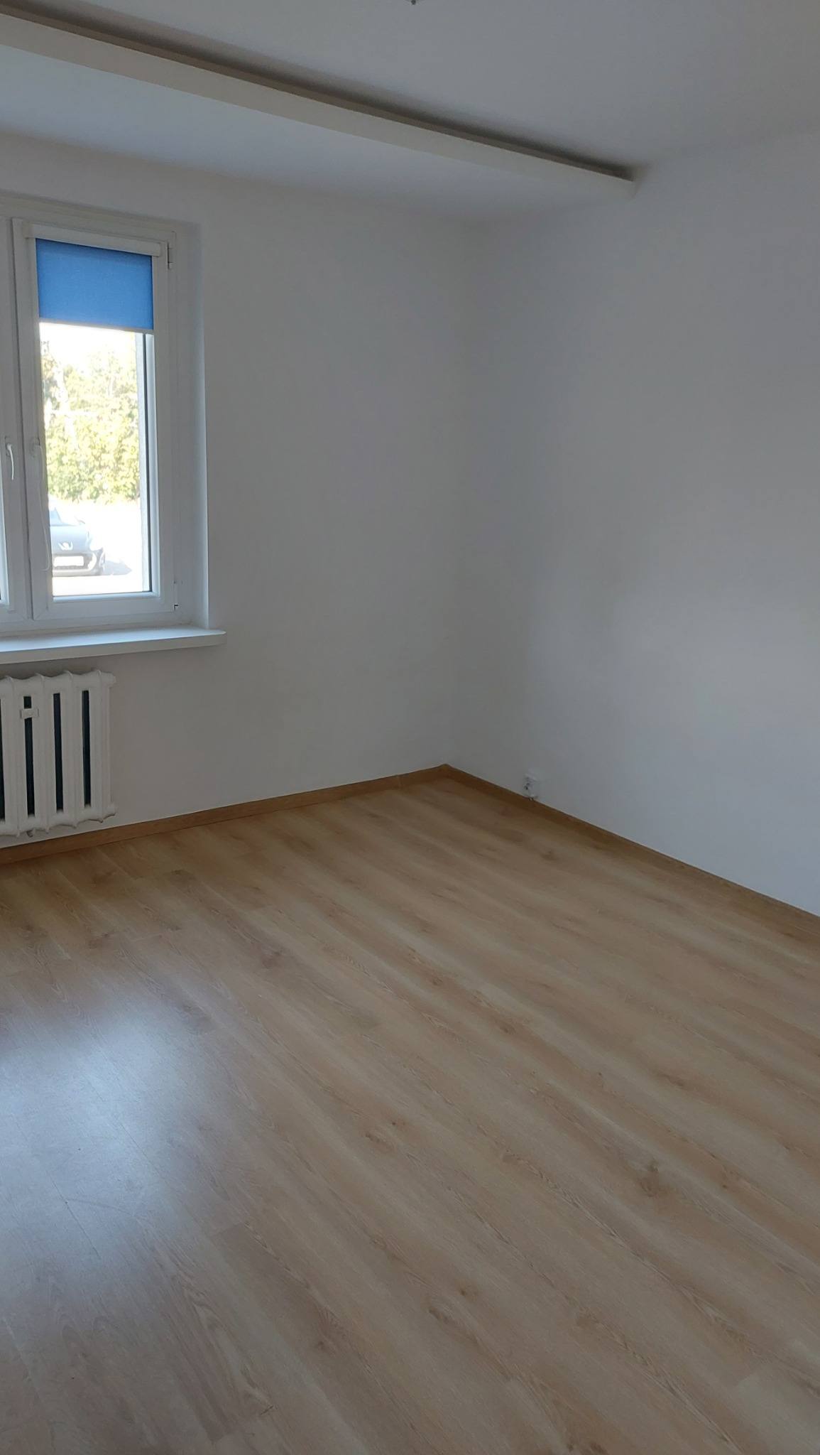 Pusty pokój po remoncie: panele podłogowe w odcieniach beżu, białe ściany, grzejnik żeberkowy pod oknem z niebieską roletą.